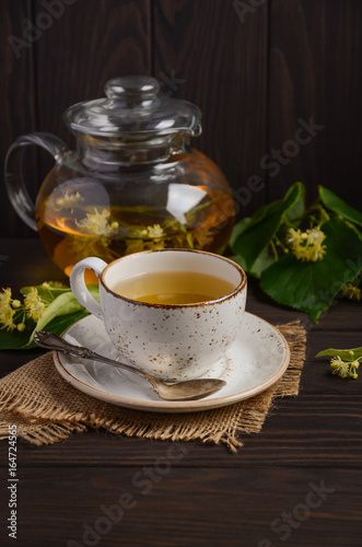 Teapot and cup with linden tea and flowers on dark wooden table, selective focus