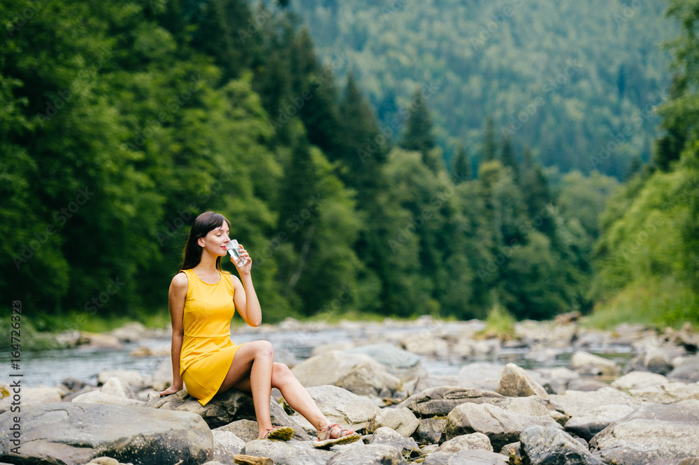 Young lonely tourist cutie girl sitting on stone on shore of river in mountains in wild terrain and satisfying thirst drinking water from glass. Traveler resting at nature. Fluid balance. Heat weather