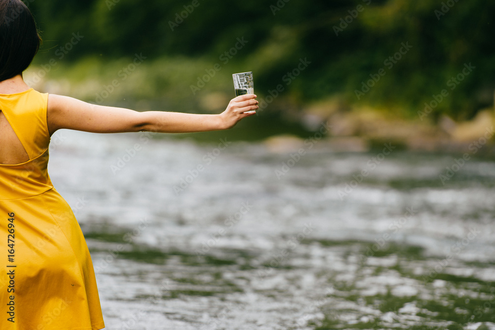 Tanned sexy young skinny girl with wet legs in yellow dress standing in  mountain river with steam. Temptation. Cutie holding glass with clean water  in hand. Thirst at nature. Proposing, advertising. Stock