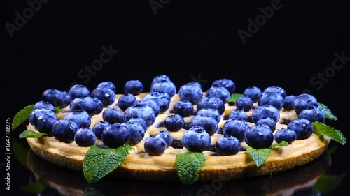 Excellent side-view of round pie with fresh blueberries and mint leaves, rotating against clock. Amazing dessert on black background with vibrant texture in 4k, 3840x2160, clip.