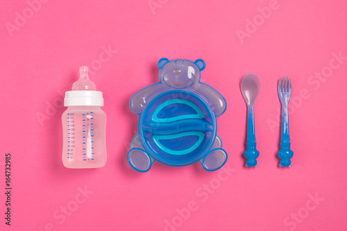 Baby bottle and blue plate with spoon and fork isolated on pink. Top view