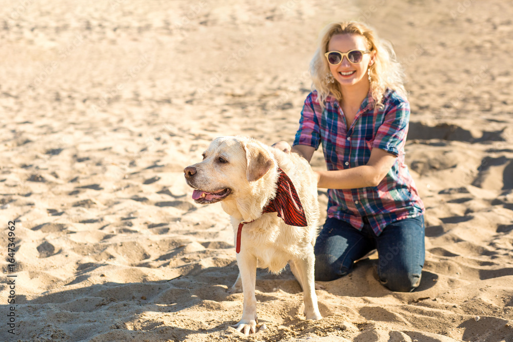 Portrait of young beautiful woman in sunglasses sitting on sand beach with golden retriever dog. Girl with dog by sea.