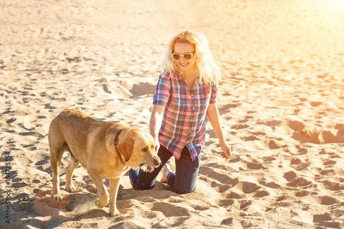 Portrait of young beautiful woman in sunglasses sitting on sand beach with golden retriever dog. Girl with dog by sea. Sun flare