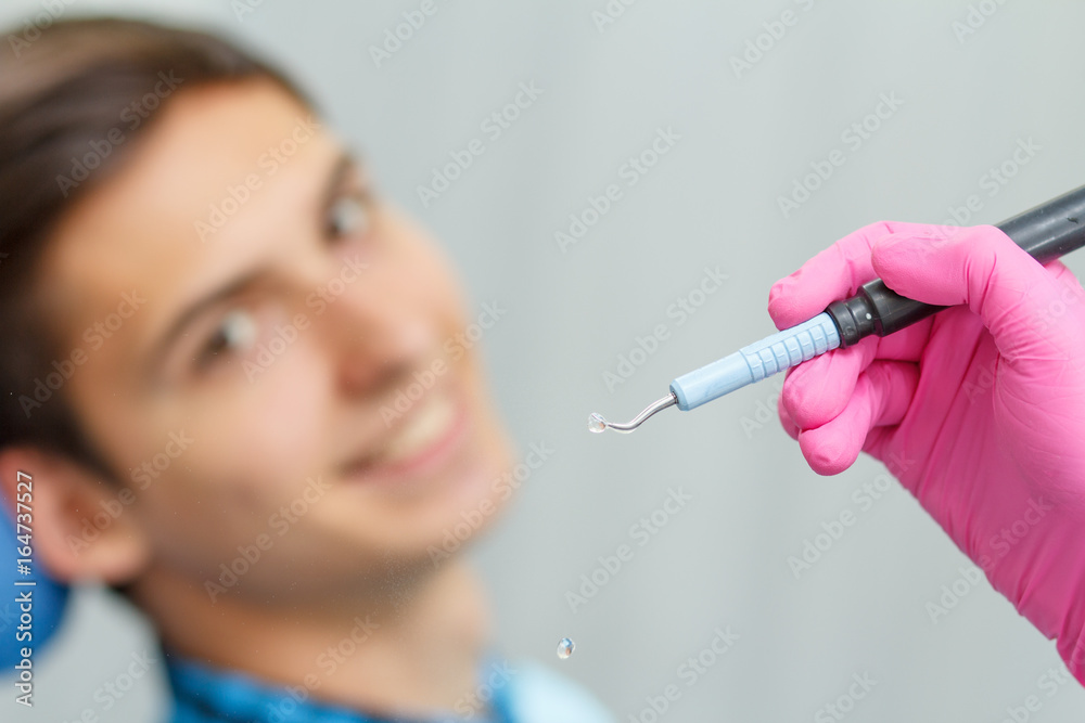 Doctor wearing pink gloves is holding ultrasonic dental scaler with young man smiling on the background Doctor wearing pink gloves is holding ultrasonic dental scaler with young man smiling on the background