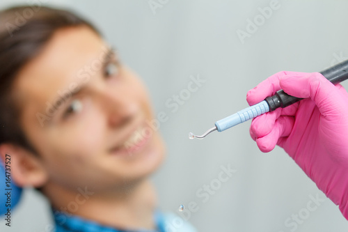 Doctor wearing pink gloves is holding ultrasonic dental scaler with young man smiling on the background