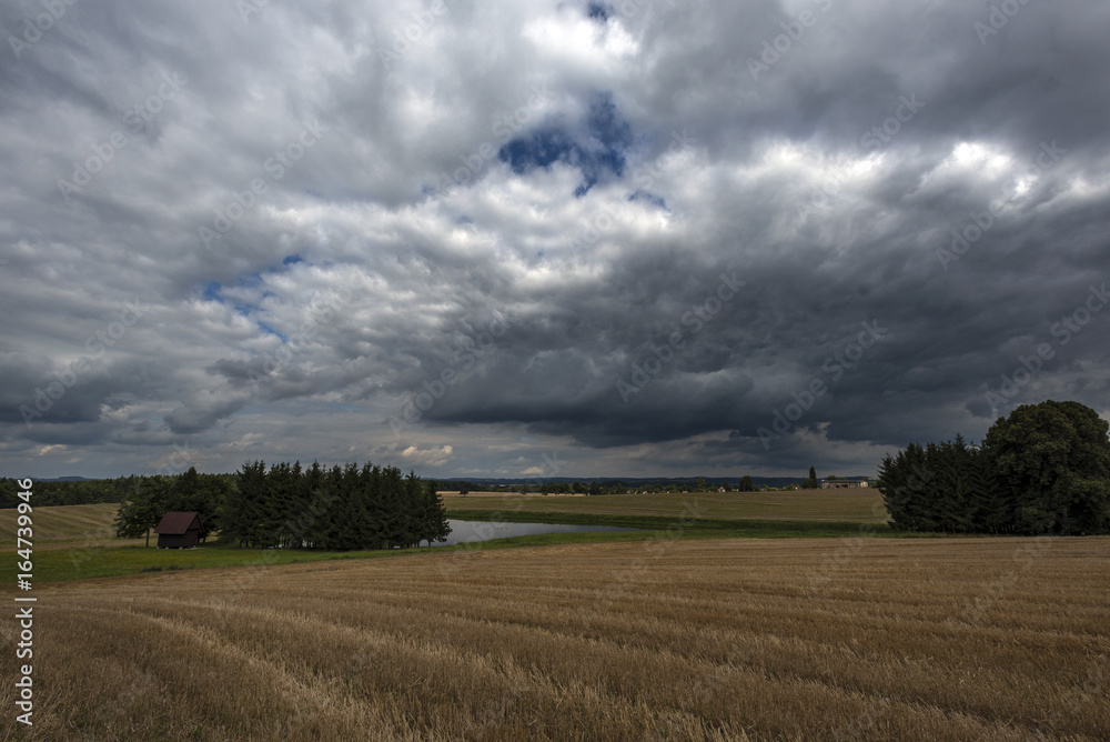 Hunting lodge near a lake near the forest, field of grain under storm clouds before sunset in the highlands in Bohemia Hunting lodge near a lake near the forest, field of grain under storm clouds before sunset in the highlands in Bohemia
