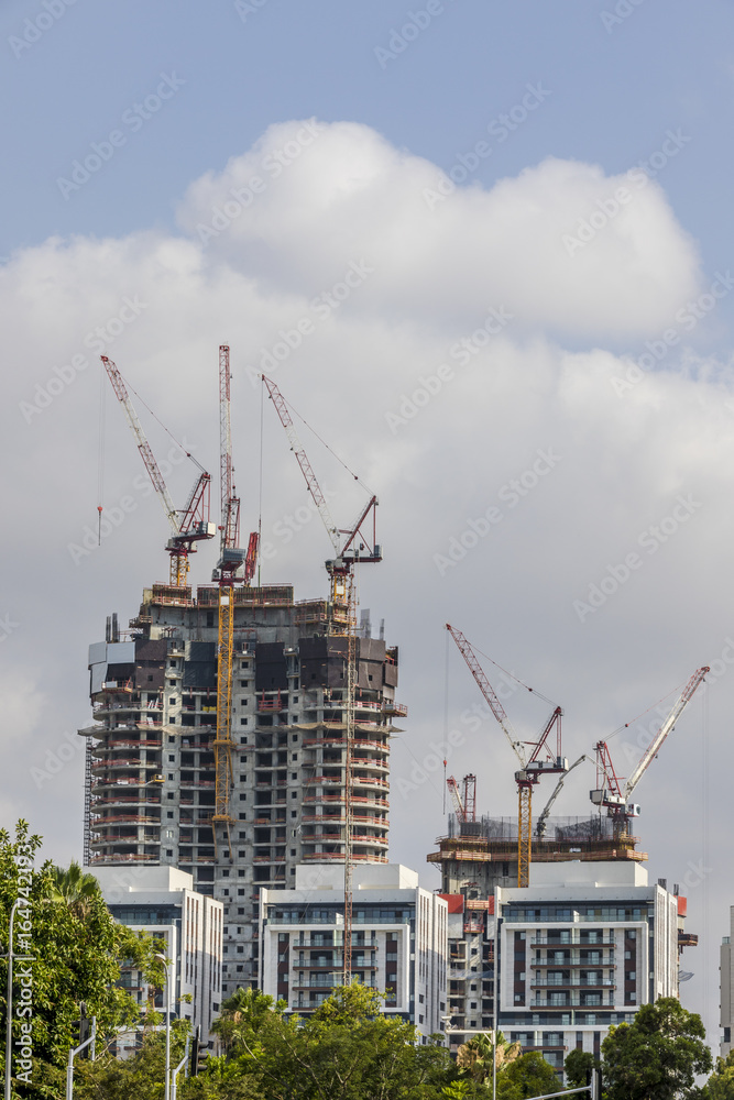 Two buildings one multistory under construction with scaffolds and cranes on top of it green tree cloudy sky behind white residential buildings