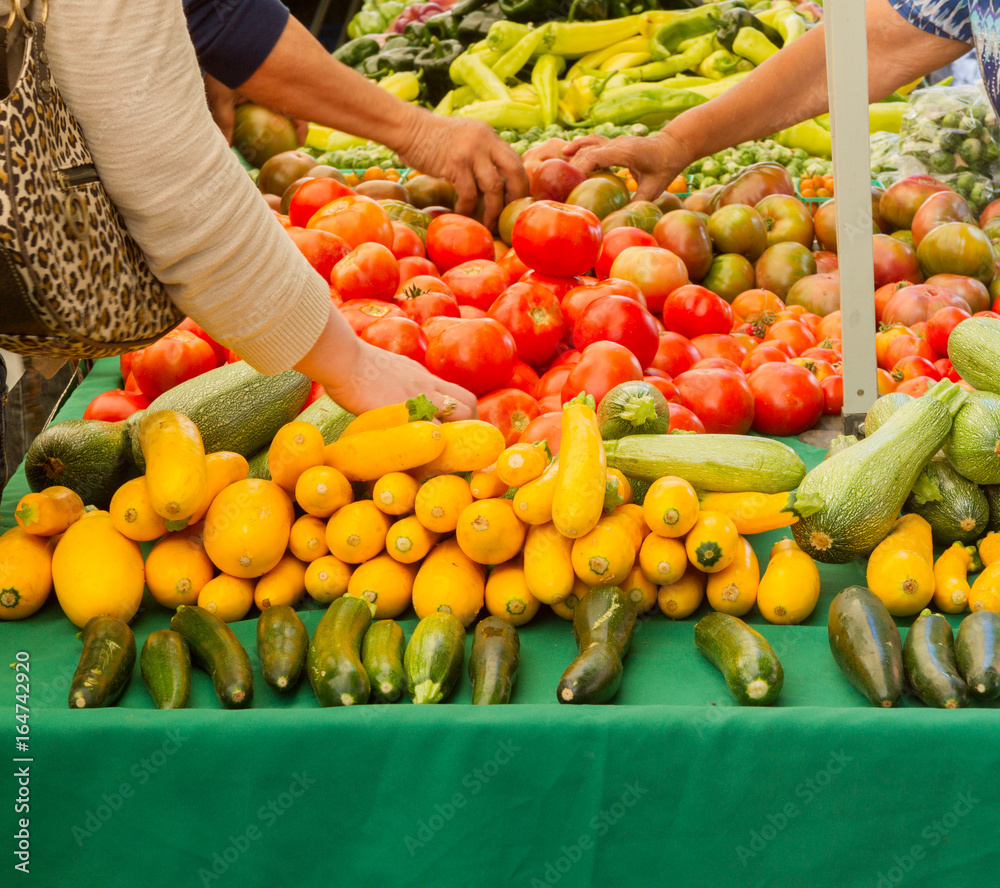 Farmer's market produce for sale at stand, with one woman reaching for a squash on left and another woman holding tomatoes on the right. Tomatoes, squashes, gypsy peppers and bell peppers are visible.
