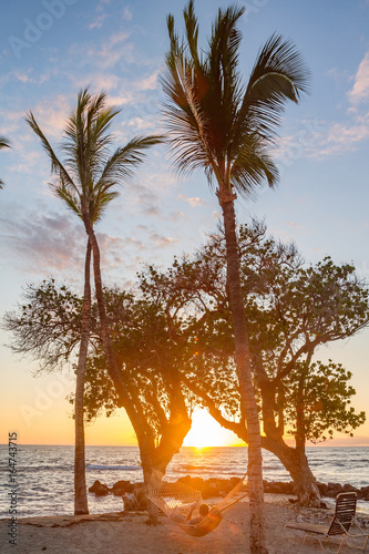 couple laying in hammock between silhouetted trees on Hawaiian beach at sunset. palm trees frame hammock with sun setting in distance. Orange, yellow and pink hues with few wispy clouds
