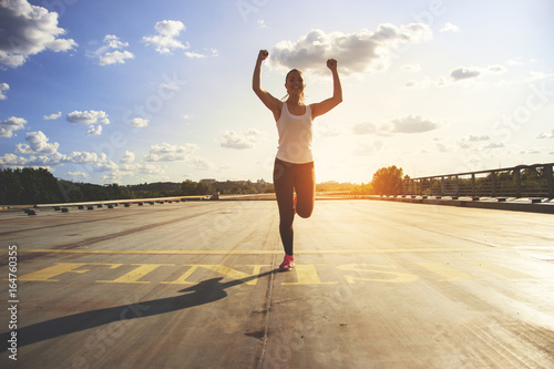 Winner as style of life. Horizontal shot of young beautiful woman in sports clothing keeping arms raised and smiling while passing finish line during jogging. Evening sunlight on background. photo