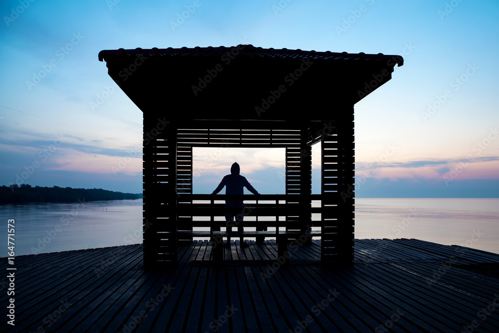 Frustrated depressed man wearing hoodie standing alone on wooden bridge extended into the sea looking down and contemplating suicide., Concept of unemployed, sadness, depression and human problems.