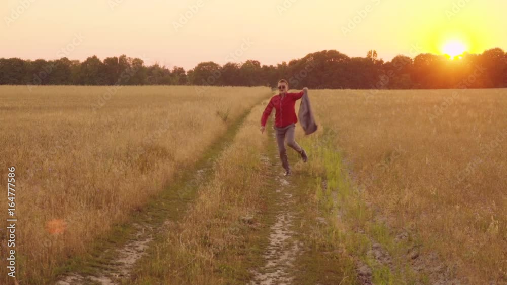 Successful businessman runs skipping along a country road near a wheat field at sunset, waving his jacket in slow motion Successful businessman runs skipping along a country road near a wheat field at sunset, waving his jacket in slow motion