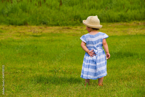 A little girl in a hat and in a blue dress is walking along the lawn © Angelina