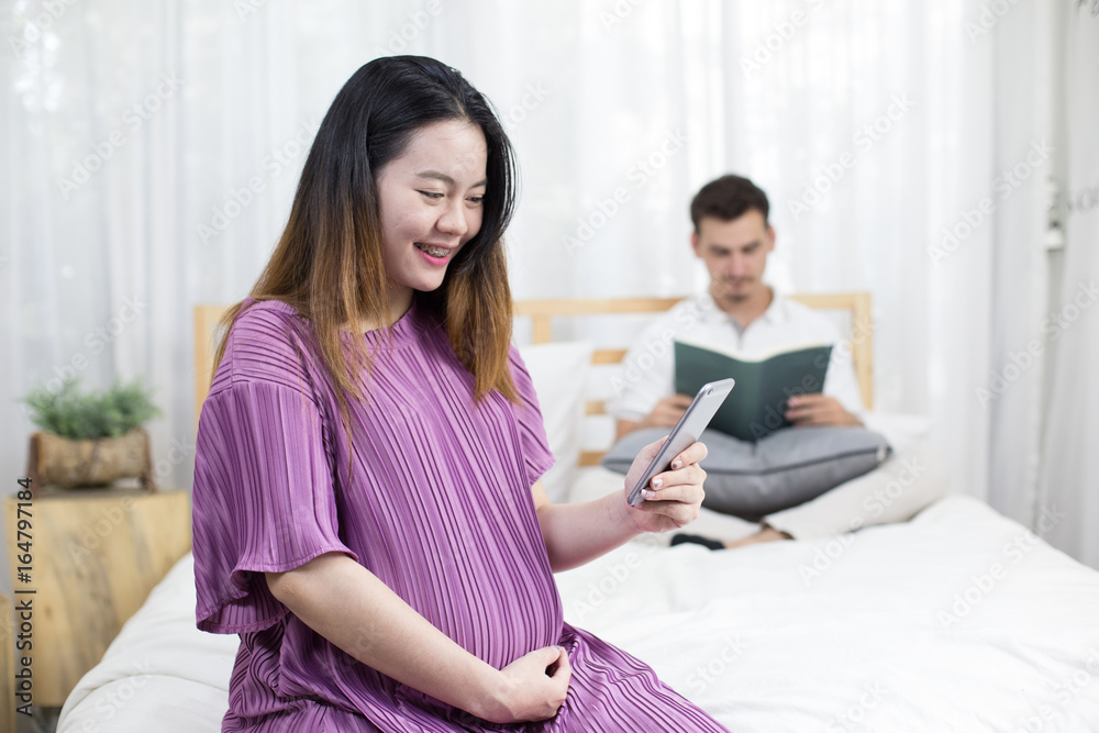 Asian pregnant woman holding smartphone and husband behind at bedroom together, 2 person, pregnant woman with happy emotion concept. Asian pregnant woman holding smartphone and husband behind at bedroom together, 2 person, pregnant woman with happy emotion concept.