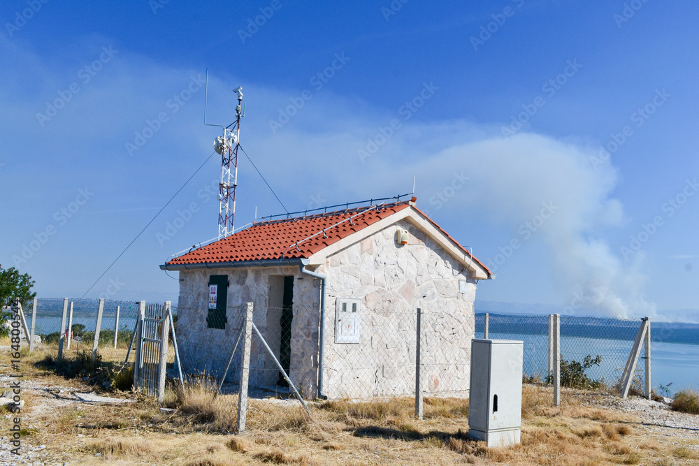 Radio telecommunication small building with red roof at the top of the mountain in Croatia, Dalmatia. In the background Vransko lake and white smoke.