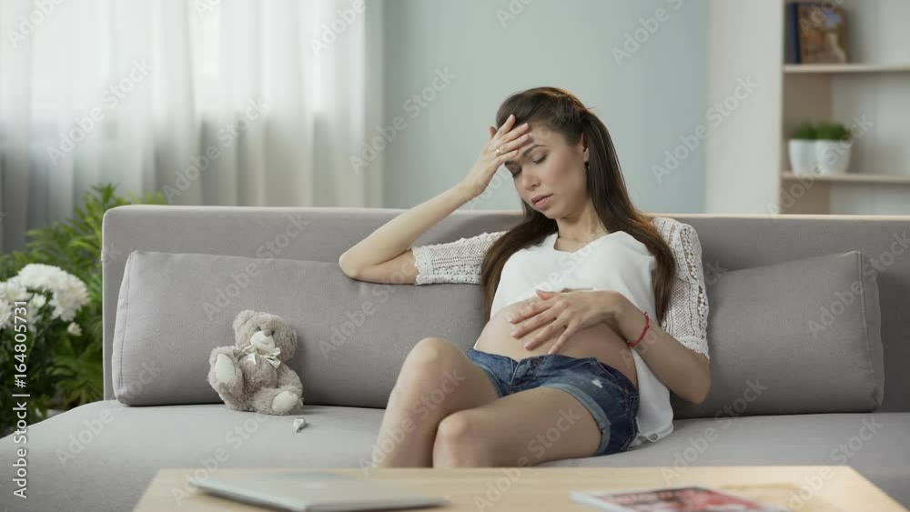 Woman pregnant with child touching belly and head, looking preoccupied, worries Woman pregnant with child touching belly and head, looking preoccupied, worries