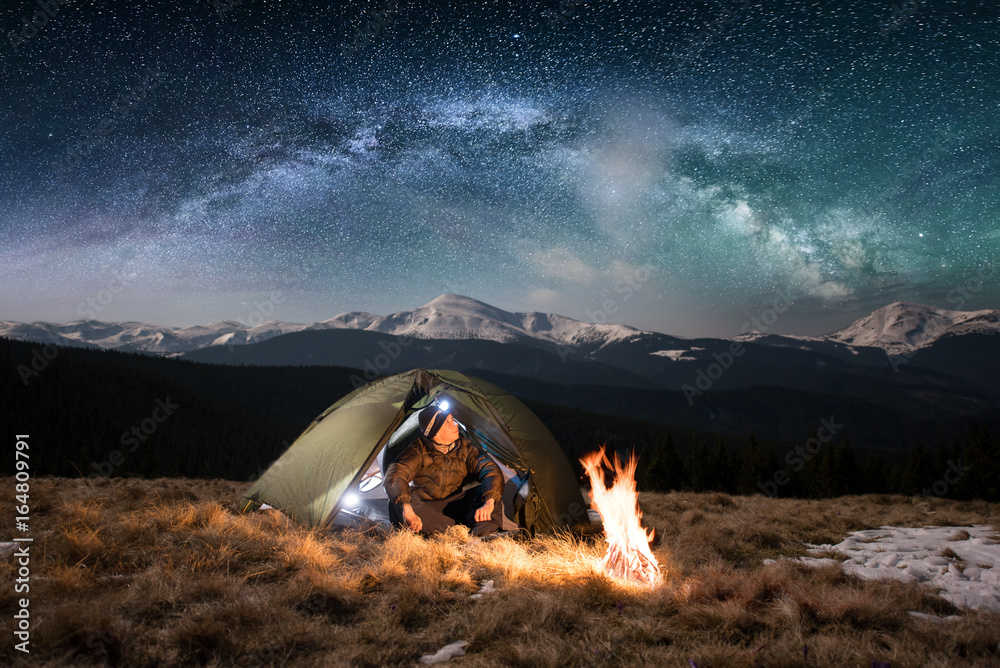 Male tourist have a rest in his camping in the mountains at night. Man with a headlamp sitting near campfire and tent under beautiful night sky full of stars and milky way, and enjoying night scene - obrazy, fototapety, plakaty 
