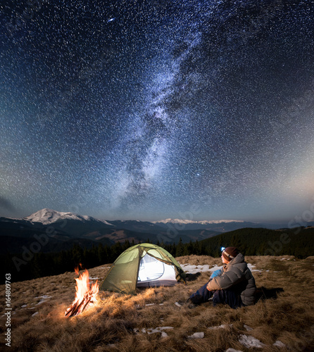 Male tourist have a rest in his camp at night. Man with a headlamp sitting near campfire and tent under beautiful sky full of stars and milky way. On the background snow-covered mountains and forests