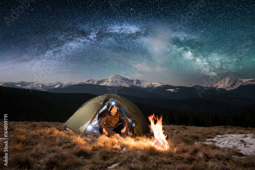 Male tourist have a rest in his camping in the mountains at night. Man with a headlamp sitting near campfire and tent under beautiful night sky full of stars and milky way, and enjoying night scene