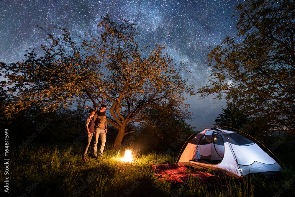 Romantic pair tourists standing at a campfire near tent under trees and beautiful night sky full of stars and milky way. Night camping. Astrophotography - obrazy, fototapety, plakaty 