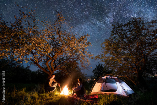 Romantic couple hikers sitting at a campfire near tent under trees and beautiful night sky full of stars and milky way. Night camping