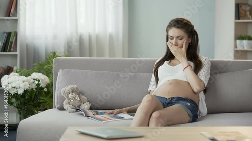 Mother-to-be sitting on couch and reading magazine, having disturbing hiccups