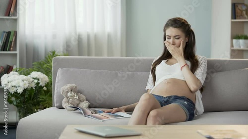 Mother-to-be sitting on couch and reading magazine, having disturbing hiccups photo