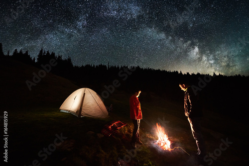 Night camping. Couple tourists standing at a campfire near illuminated tent under beautiful night sky full of stars and milky way. Long exposure