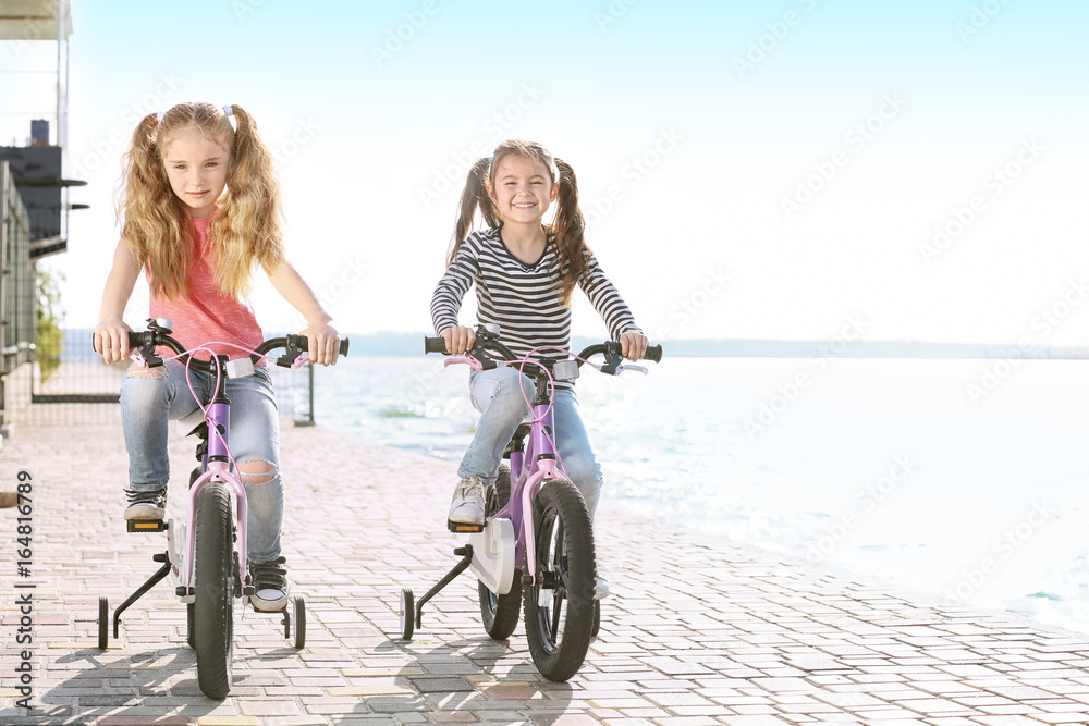 Fototapeta premium Cute little girls riding bicycles near river on sunny day