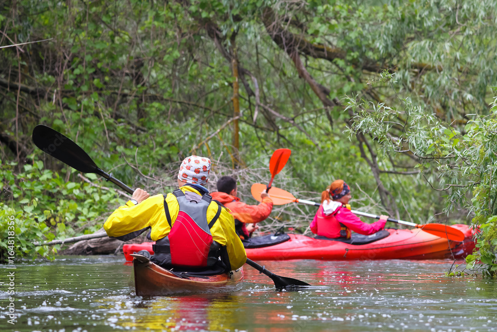 Man in selfmade brown wooden kayak in red and yellow life jacket and friends kayaking in wild river among thickets of plants on biosphere reserve in spring