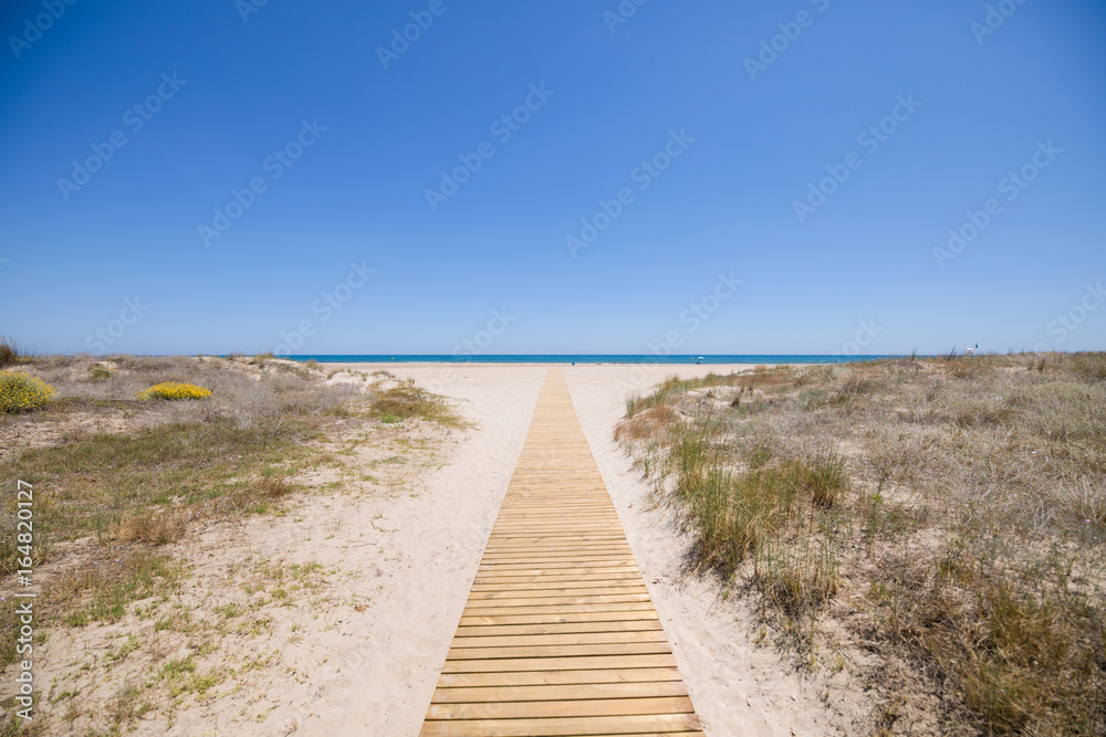 landscape access to idyllic Beach of PIne or Pinar, with wooden footway on sand and bush, in Grao of Castellon, Valencia, Spain, Europe. Blue clear sky and Mediterranean Sea 
