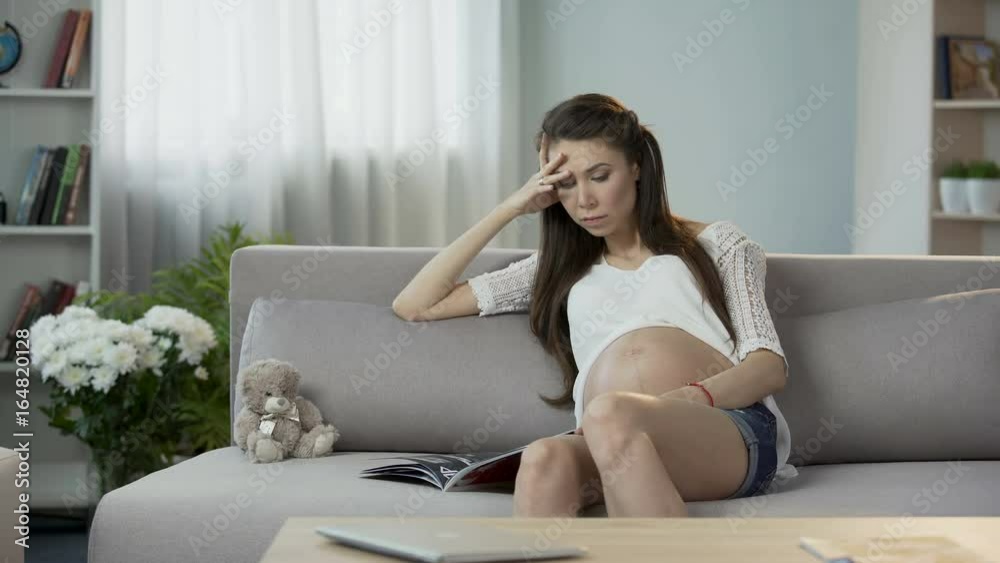 Young pregnant female sitting on sofa, reading magazine, relaxing leisure time Young pregnant female sitting on sofa, reading magazine, relaxing leisure time