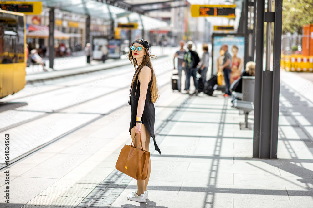 Lifestyle portrait of a stylish woman in black dress and hat walking with bag on the tram stop in the modern city