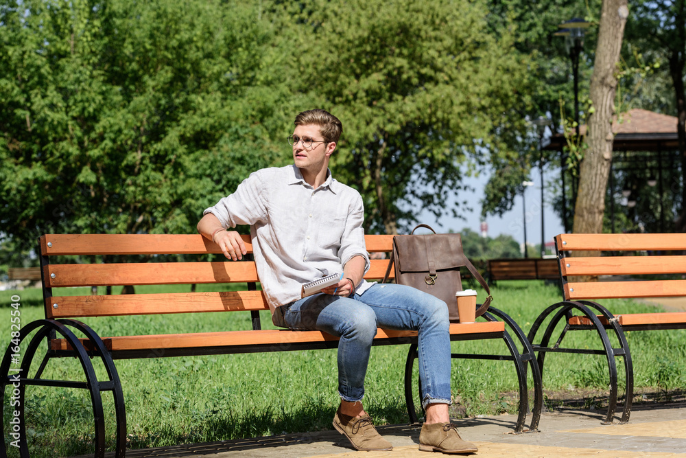 young stylish student with notebook resting on bench in park