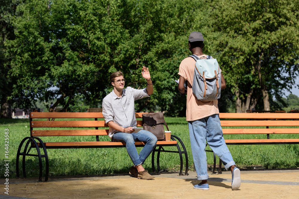 smiling man sitting on bench and waving to friend in park