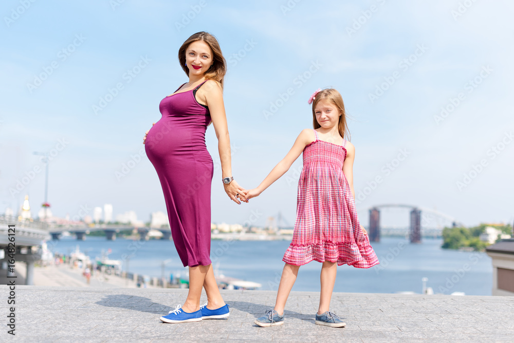 Happy family, pregnant mother and her daughter little girl child walking and hugging on the embankment in the summer day.