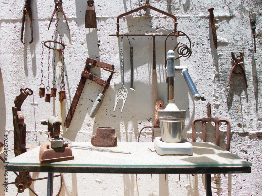 Collection of antique tools hanging on a white concrete wall and on a table in the sunshine