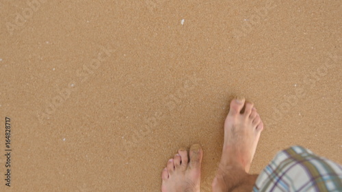 Point of view of young man stepping at the golden sand at sea beach. Male legs walking near ocean. Bare foot of guy going on sandy shore with waves. Summer vacation or holiday Close up POV
