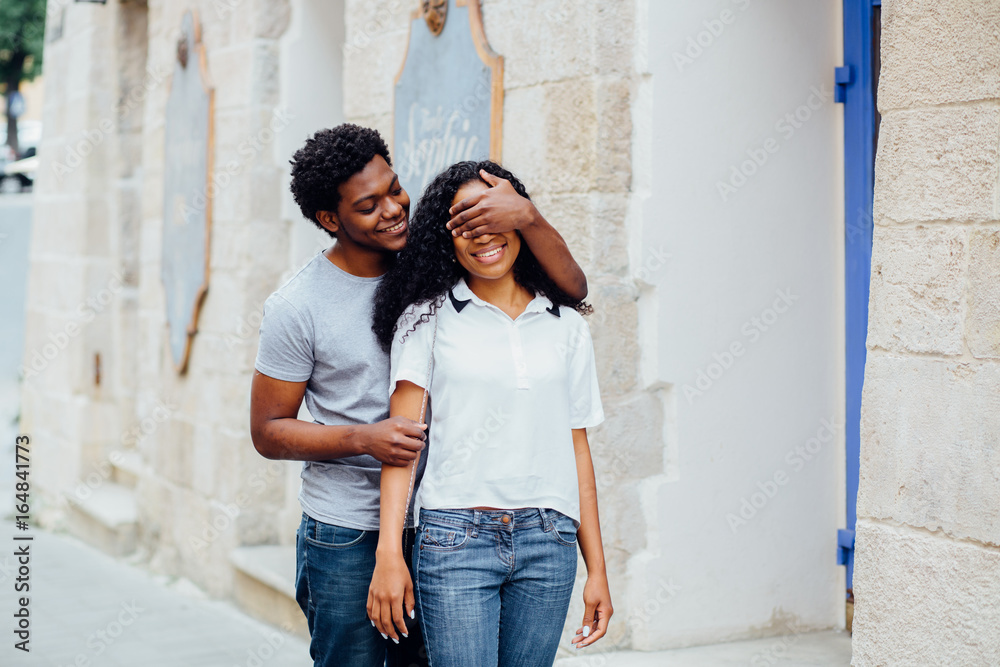 Beautiful dark skin woman smilling and trying to guess who is standing behind her. Her afro american boyfriend is covering her eyes by hands and smiling. Love and gift concept