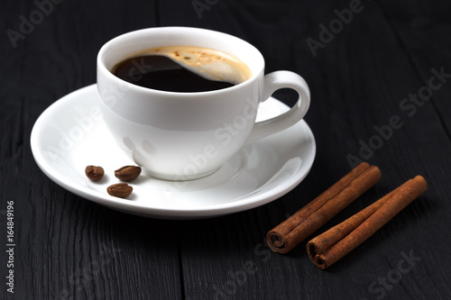 Black coffee in a white cup with cinnamon and foam on a black wooden background