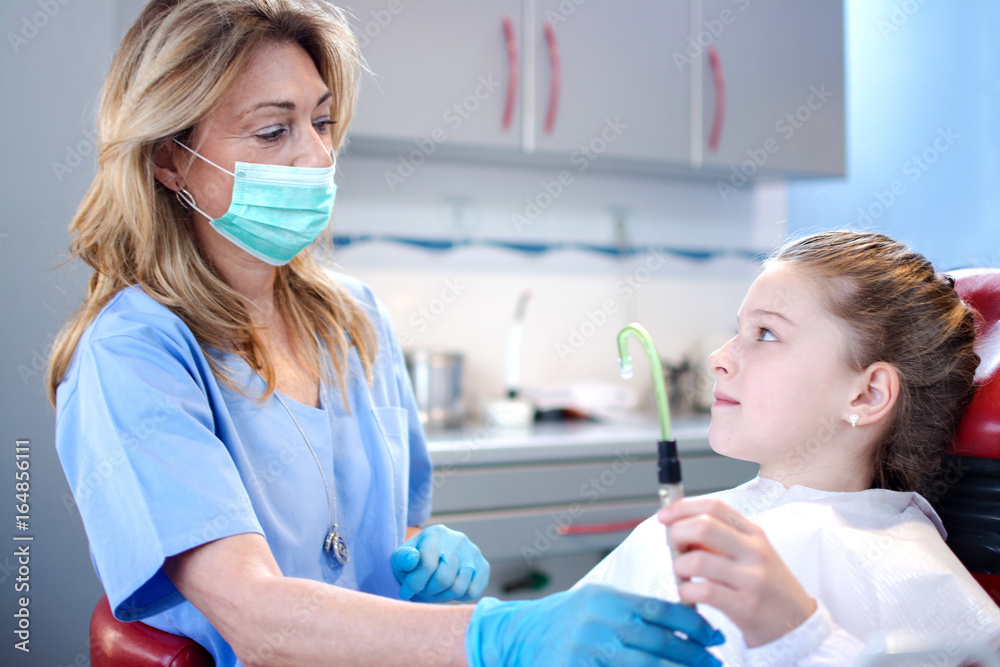 Girl patient holding suction tube near professional female dentist in dental office. Girl patient holding suction tube near professional female dentist in dental office.
