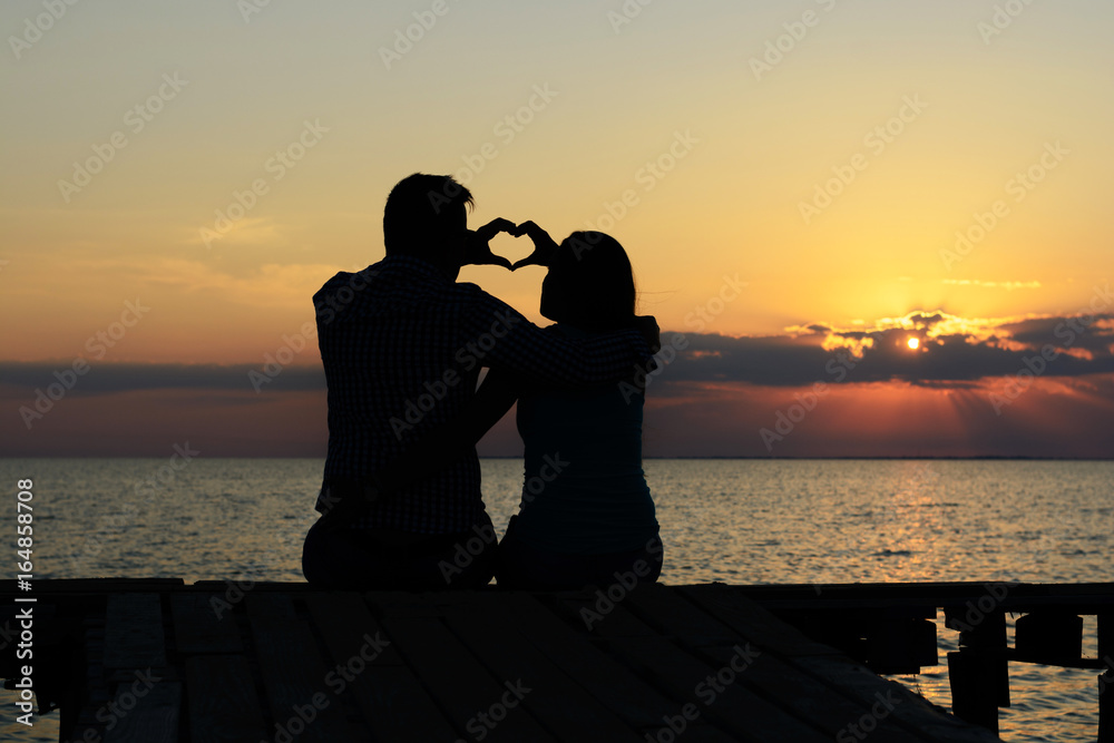 Young loving guy and girl sit on the bridge by the sea and skip a silhouette of the heart in the sky at sunset sun and sun rays. Family happy couple. Love and romance