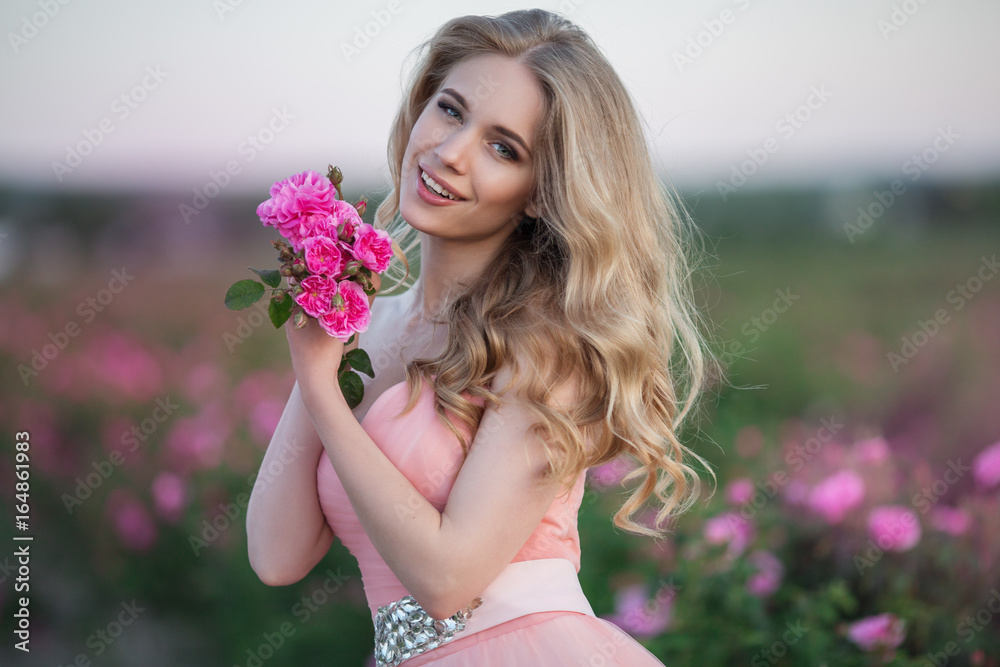 Fototapeta premium Beautiful pretty woman is wearing fashion dress posing near pink roses in a garden