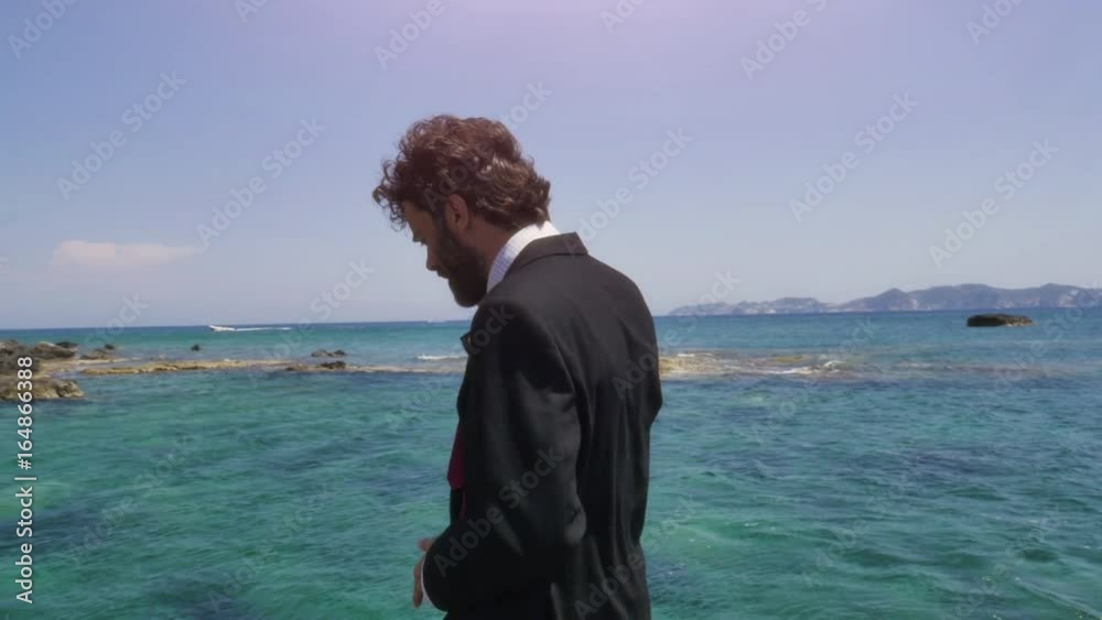 Man in suit talking on his smartphone on a deserted island in front of the ocean Man in suit talking on his smartphone on a deserted island in front of the ocean