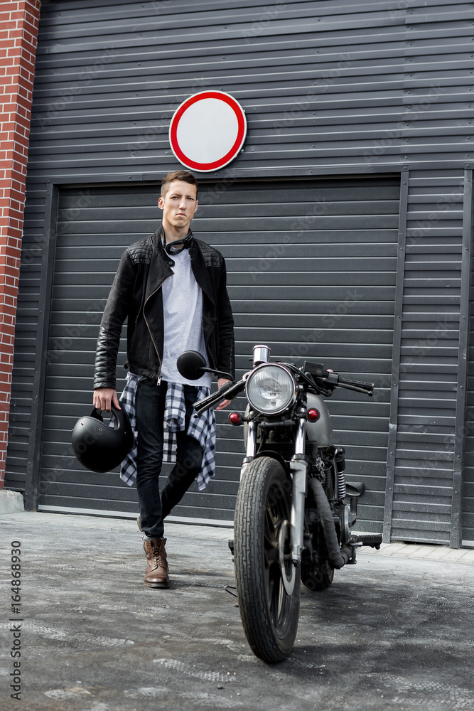 Fotka „Beautiful young rider guy in black biker jacket and boots go to his  classic style cafe racer motorcycle industrial gates as background. Bike  custom made in vintage garage. Brutal fun urban