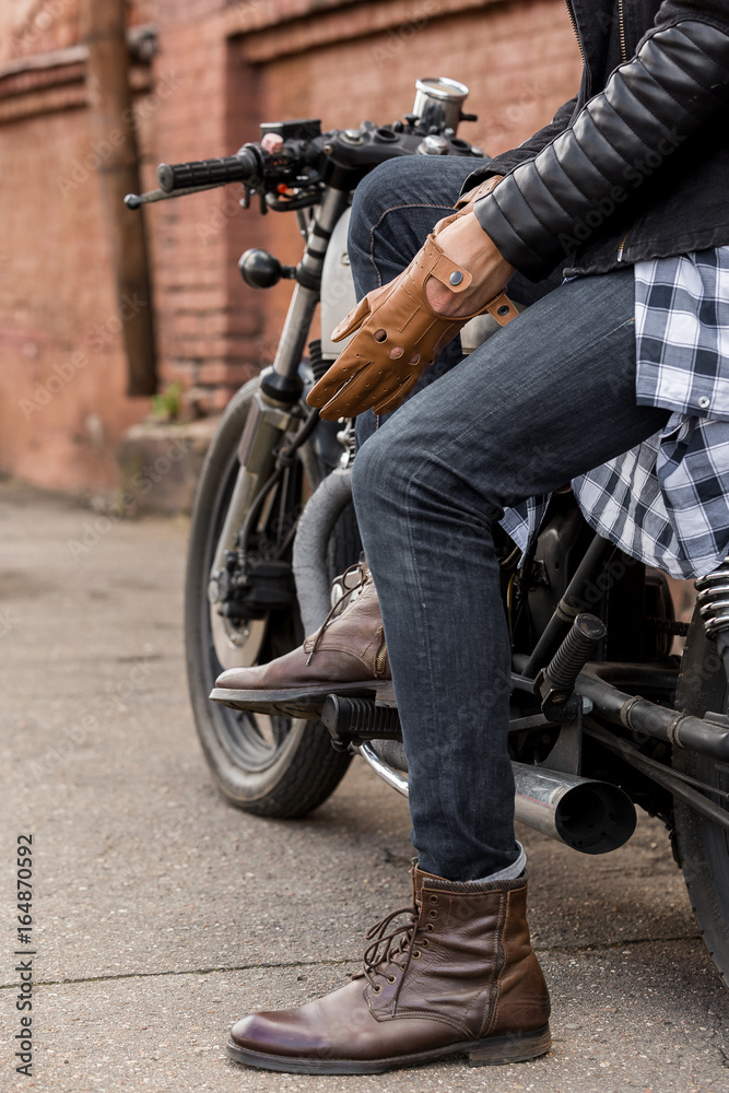 Obraz premium Close-up of a handsome rider biker man hand puts on a leather glove while sit on classic style cafe racer motorcycle. Bike custom made in vintage garage. Brutal fun urban lifestyle. Outdoor portrait.