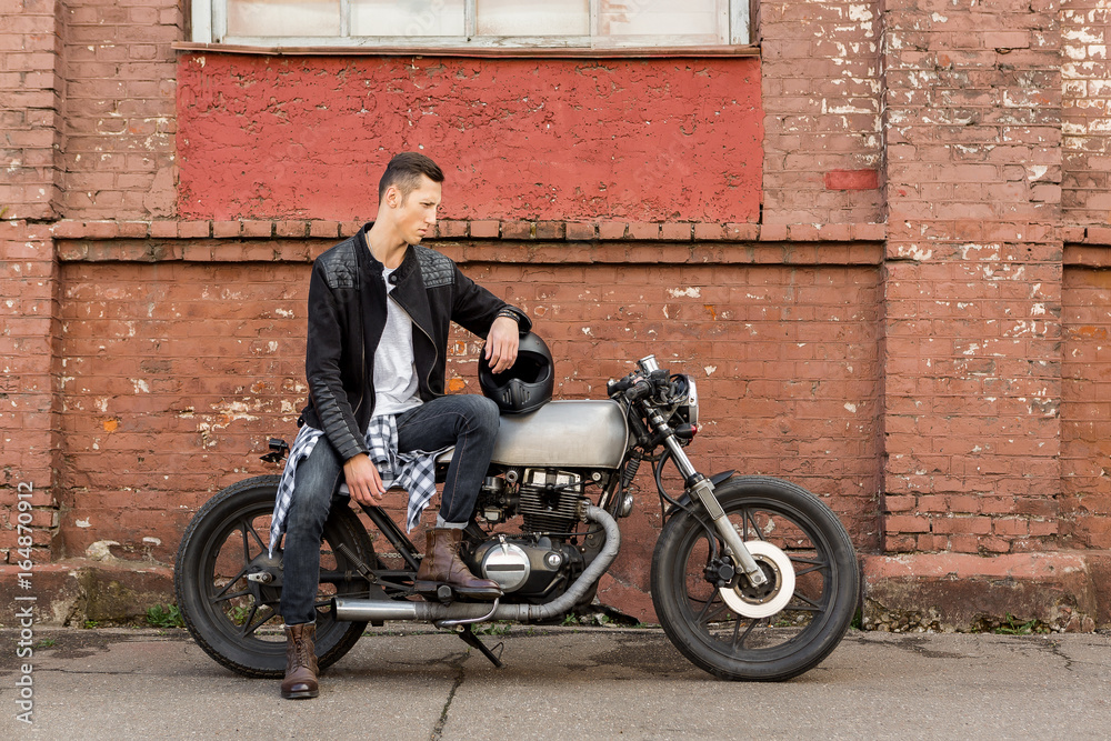 Handsome rider biker guy in black leather jacket, boots and style jeans sit  on classic style cafe racer motorcycle. Bike custom made in vintage garage.  Brutal fun urban lifestyle. Outdoor portrait. Stock