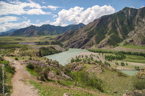 Scenic view of the winding mountain river, green fields and mountains on a background of colorful blue sky with clouds. Place of confluence of rivers Katun and Chuya. Nature and travel. Russia, Altai