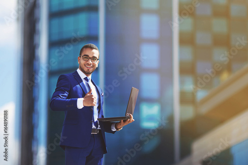 Competent business expert. Confident young handsome man in suit holding laptop and smiling while standing against office building