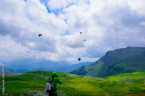 A guy is photographing black birds flying in the mountains, Alps, Italy. The landscape is represented by mountains and green meadows in a cloudy day